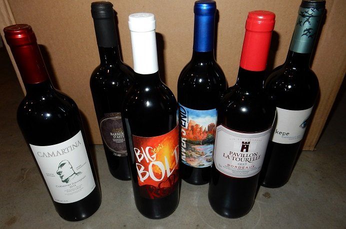 Six unopened wine bottles with different labels in front of a cardboard box