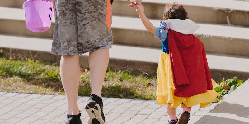 A child dressed as Snow White holds the hand of a person in shorts who is carrying a pink Halloween bucket