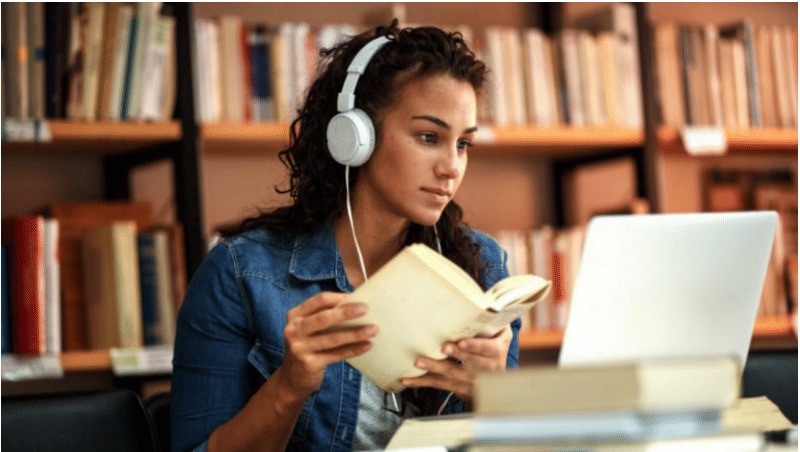 girl with headphones holding open book and looking at open laptop