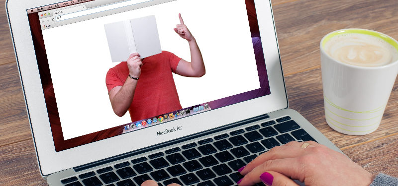 women's hands on laptop keyboard with man holding a white book in front of his face on the screen