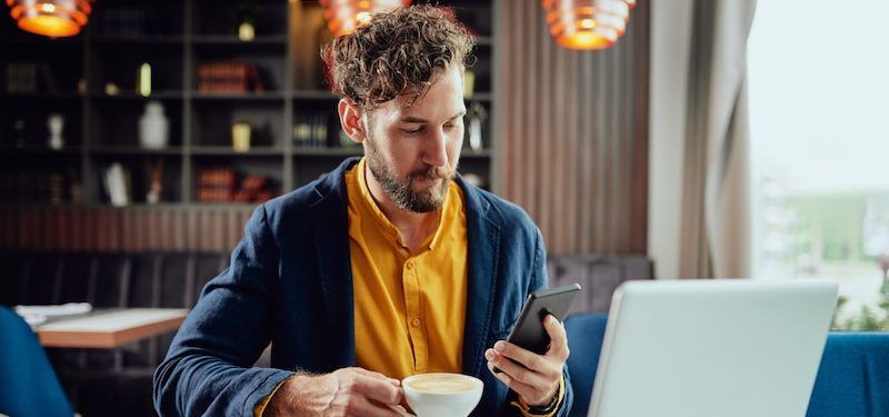 Man at coffee shop using mobile device and laptop while drinking coffee