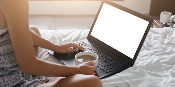 Woman sits in bed and uses computer while drinking coffee